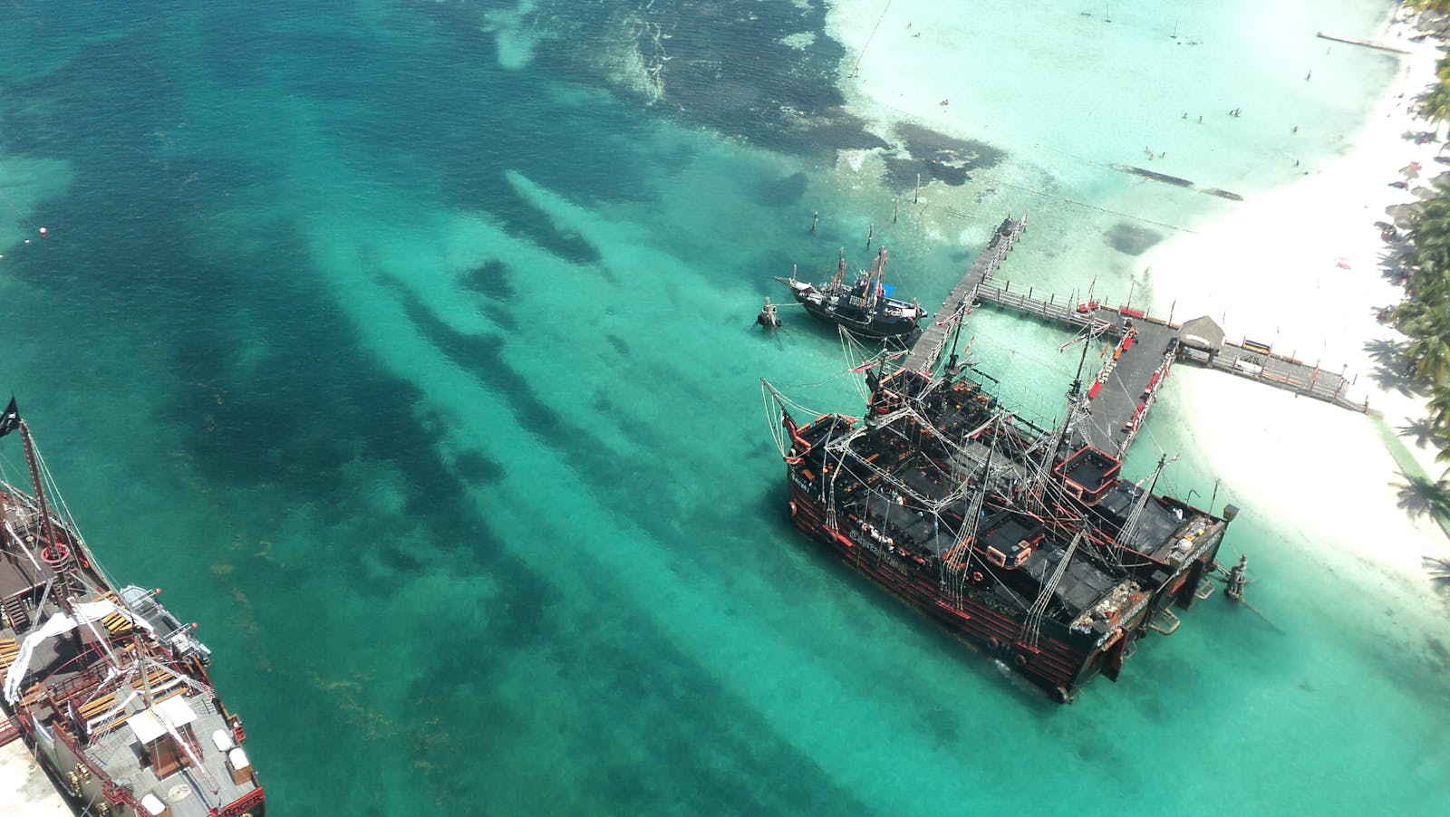 An aerial view of pirate ships near a tropical beach with clear turquoise water and white sandy shores.